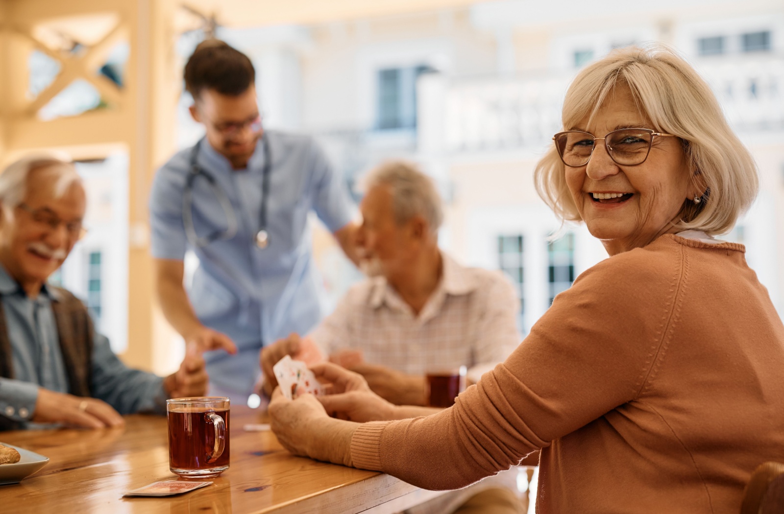 Happy older woman at a warm, sunlit communal table, playing cards with other residents at an assisted living facility.