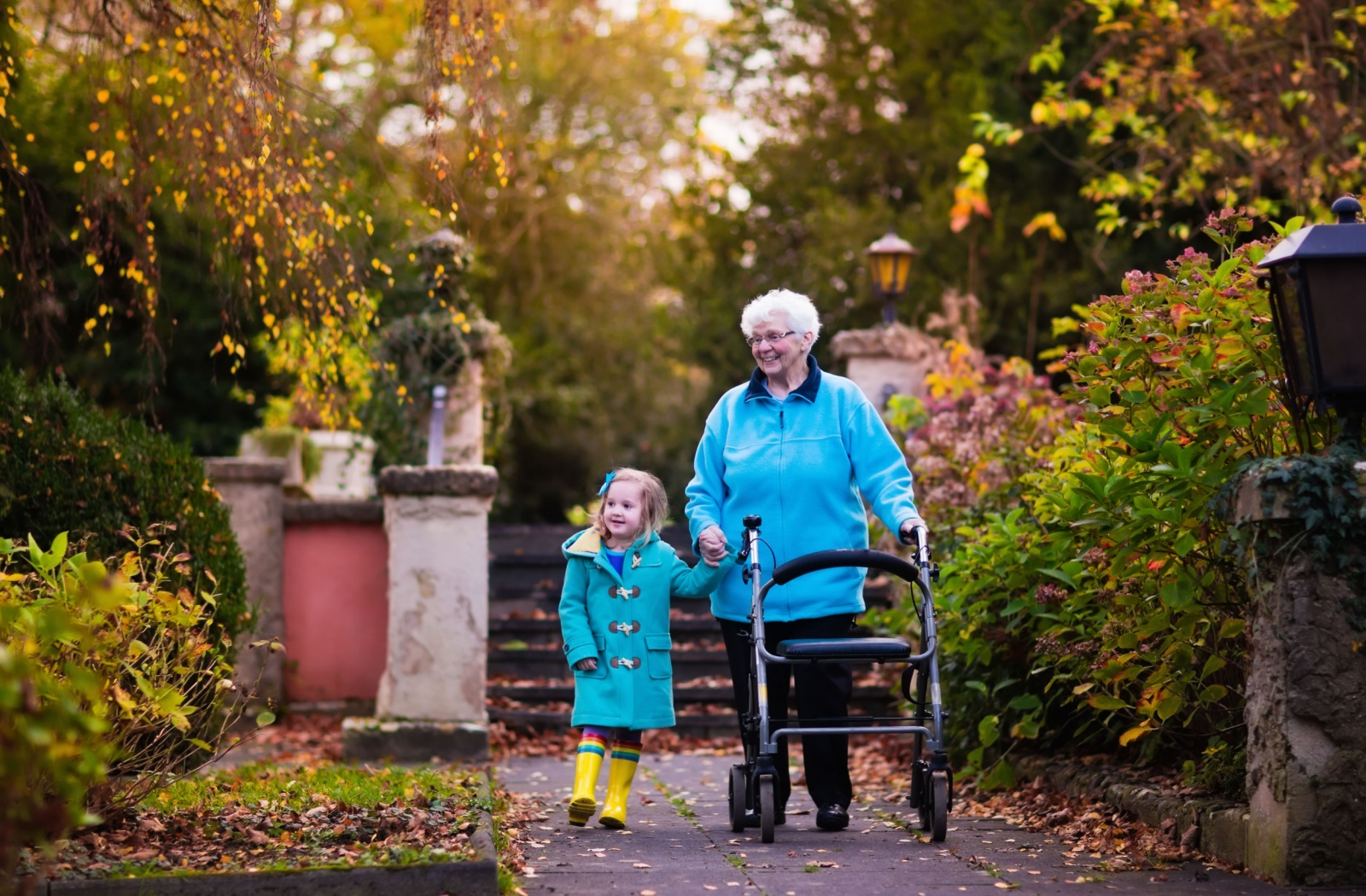 A grandparent strolls through a park with their walker and grandchild, prepared for a light rain.