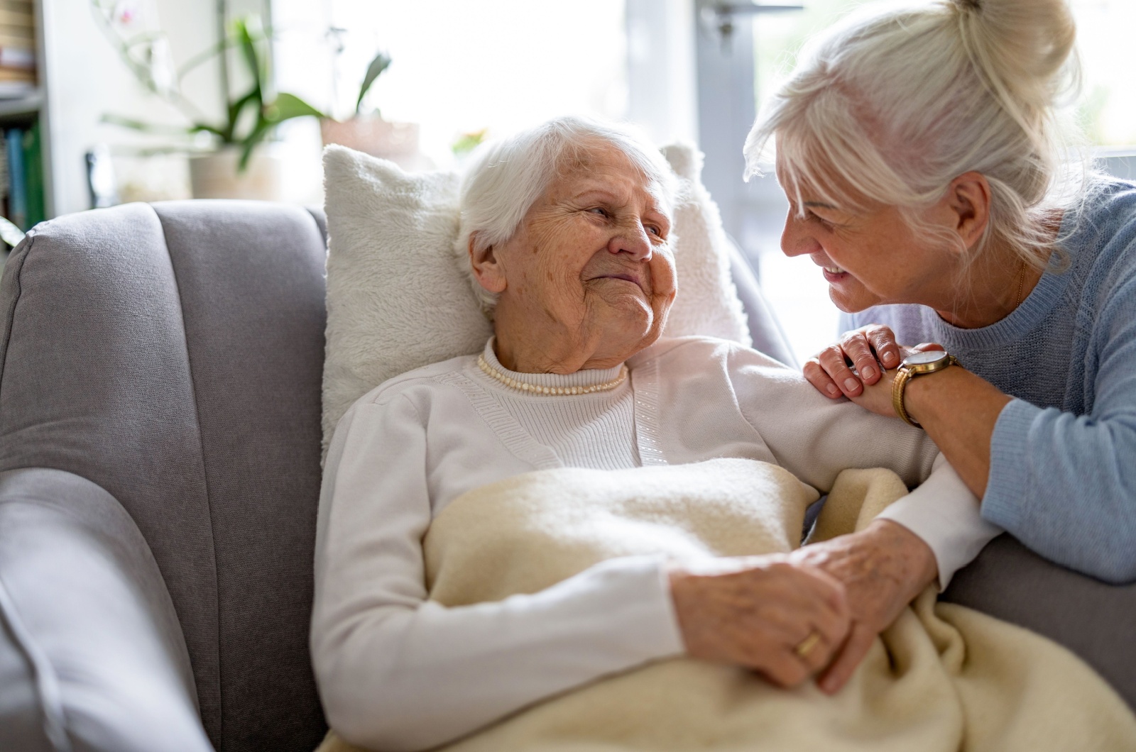 An older parent laying on a couch with a blanket, smiling at her adult child.