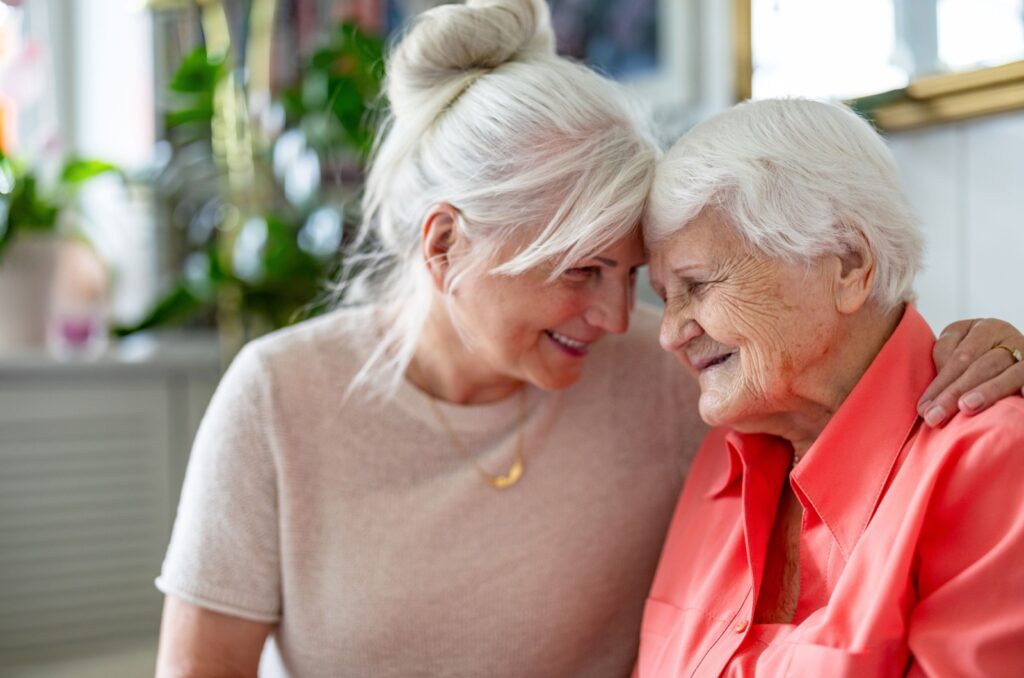 A smiling adult child discussing assisted living with her arm around her smiling older parent.