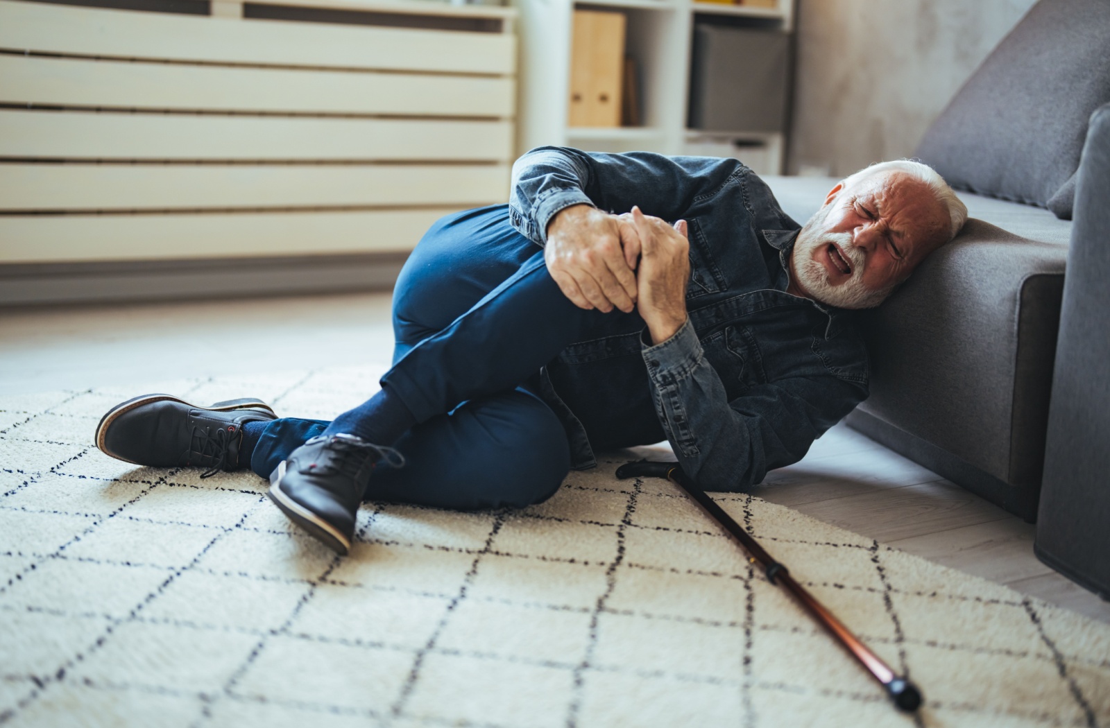 Senior in visible pain in their living room alone after a fall clenching his knee having dropped his walking cane.