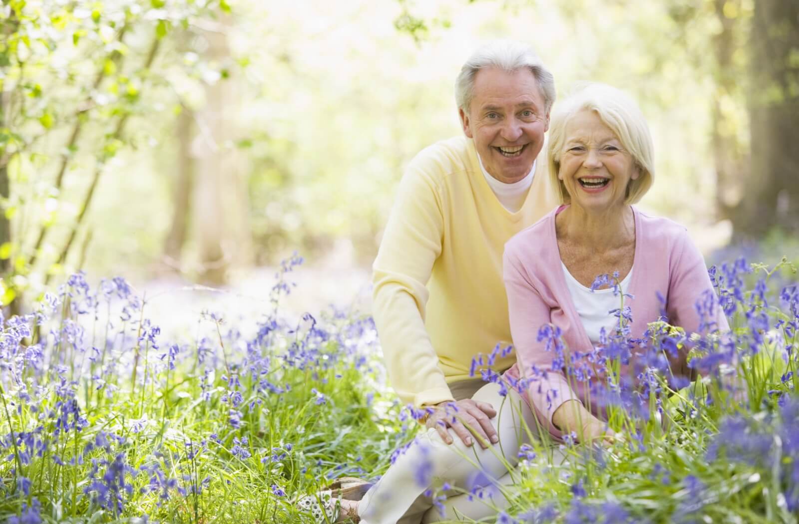 An older couple in yellow and pink shirts sitting in a field of wildflowers on a bright sunny day.