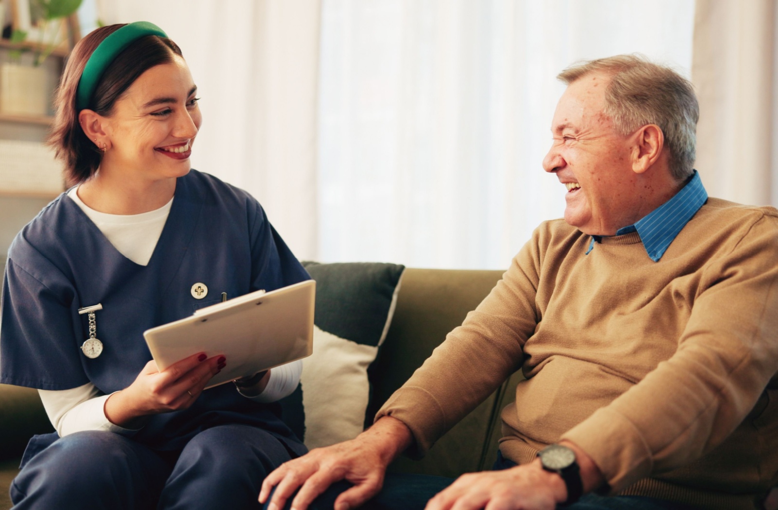 Smiling caregiver holding a clipboard while chatting with a senior seated on a couch during a wellness check-in