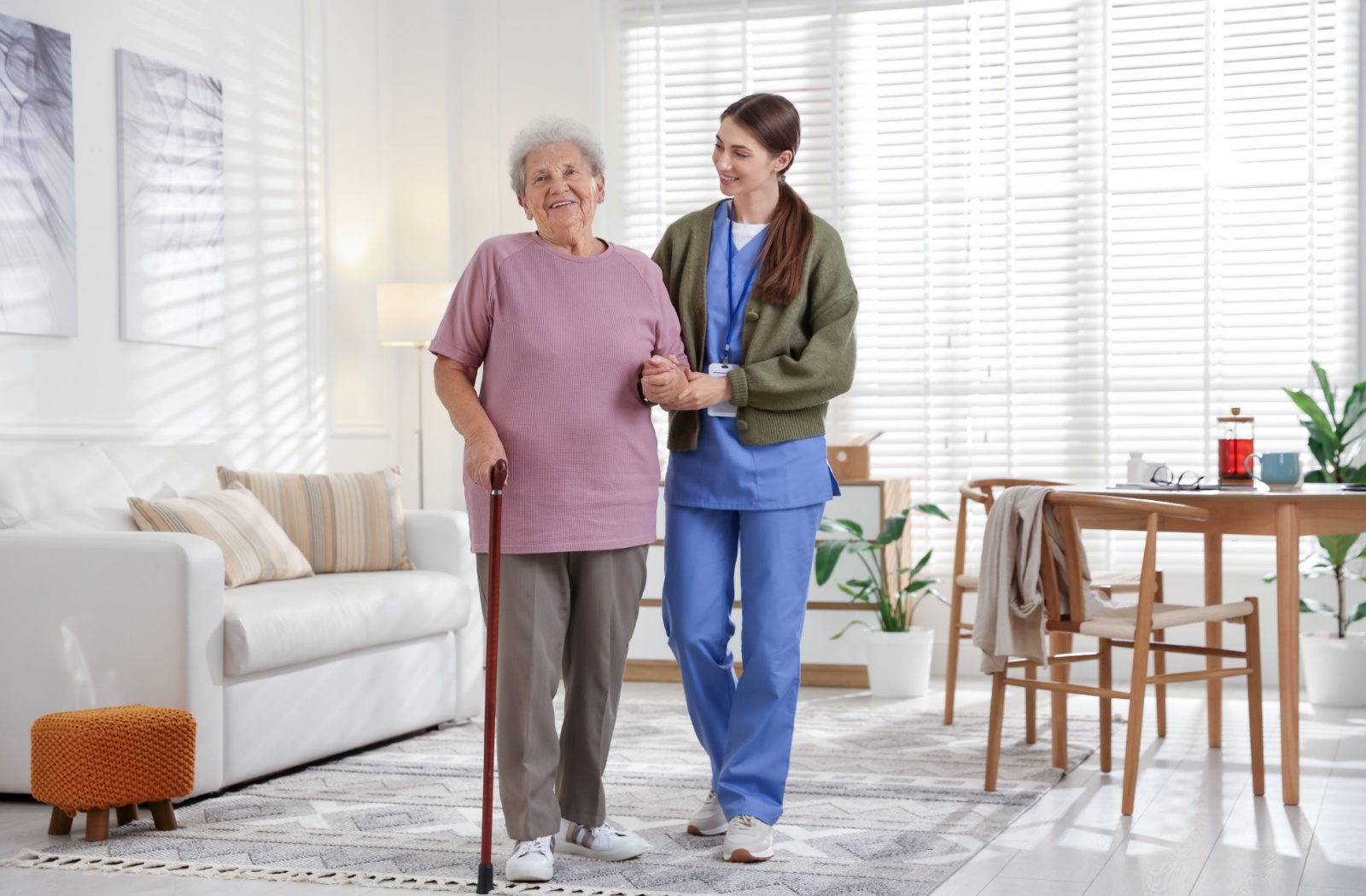 Caregiver walking with an older adult using a cane in a bright apartment to support mobility and independence.