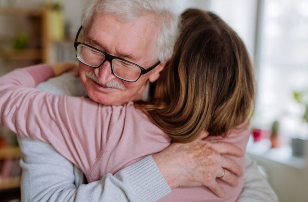 Older adult wearing glasses warmly hugging a visitor in a soft-lit room filled with natural light
