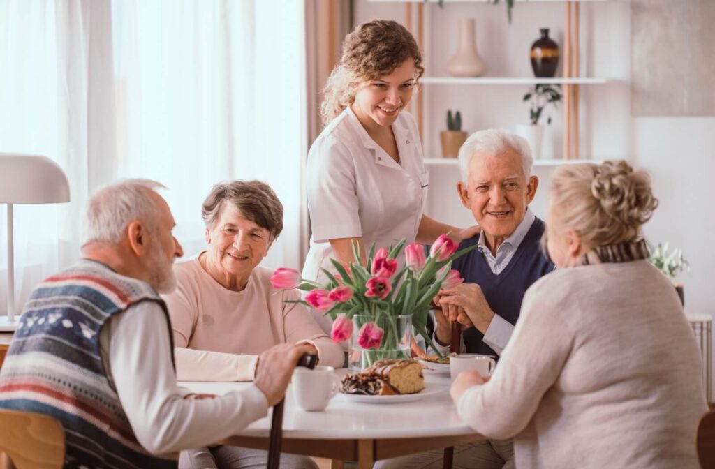 Caregiver standing beside a group of seniors enjoying coffee and cake around a table decorated with fresh pink tulips