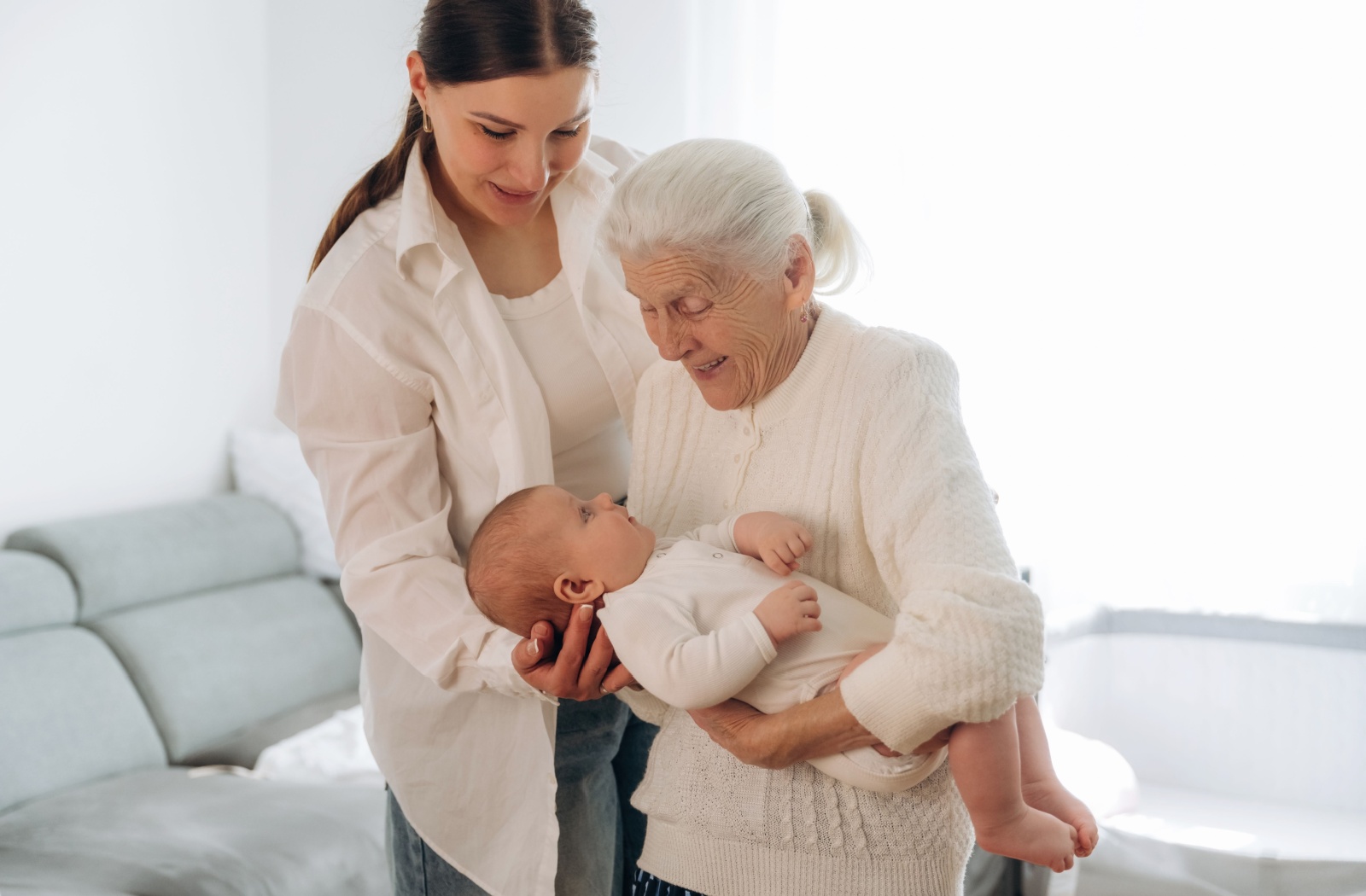 Senior holding a baby while smiling with a younger adult nearby during a multigenerational family visit
