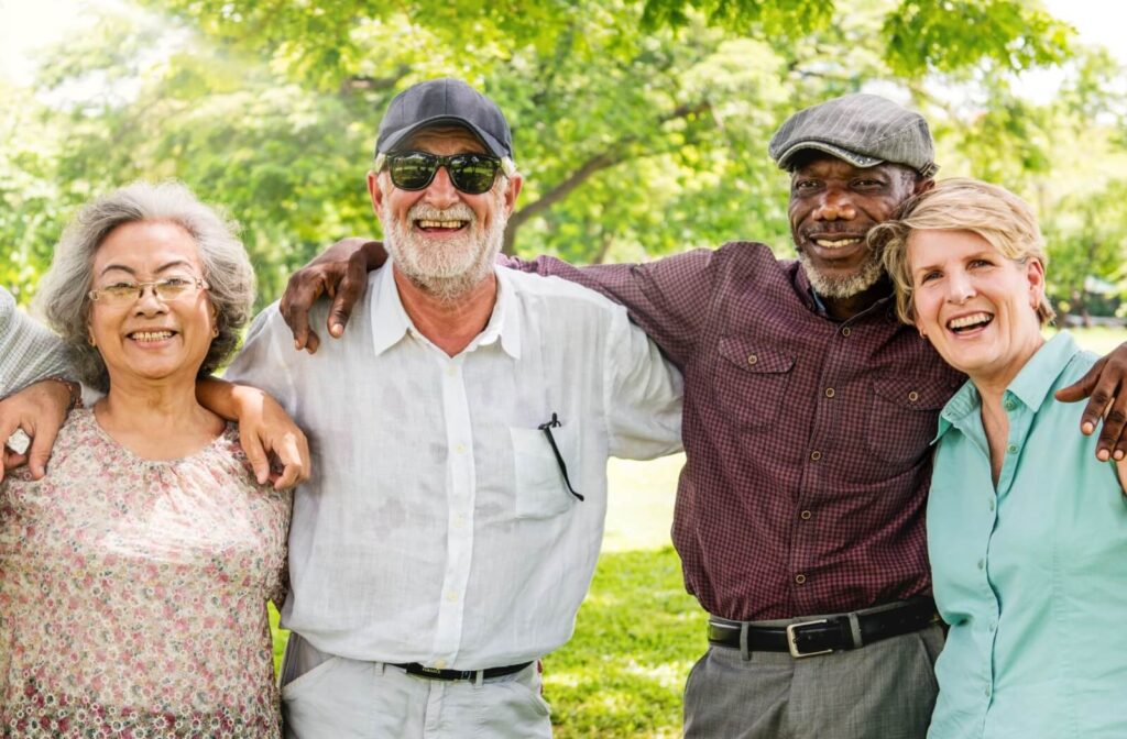 Four seniors standing arm-in-arm in a park smiling warmly with trees and green grass in the background on a bright day