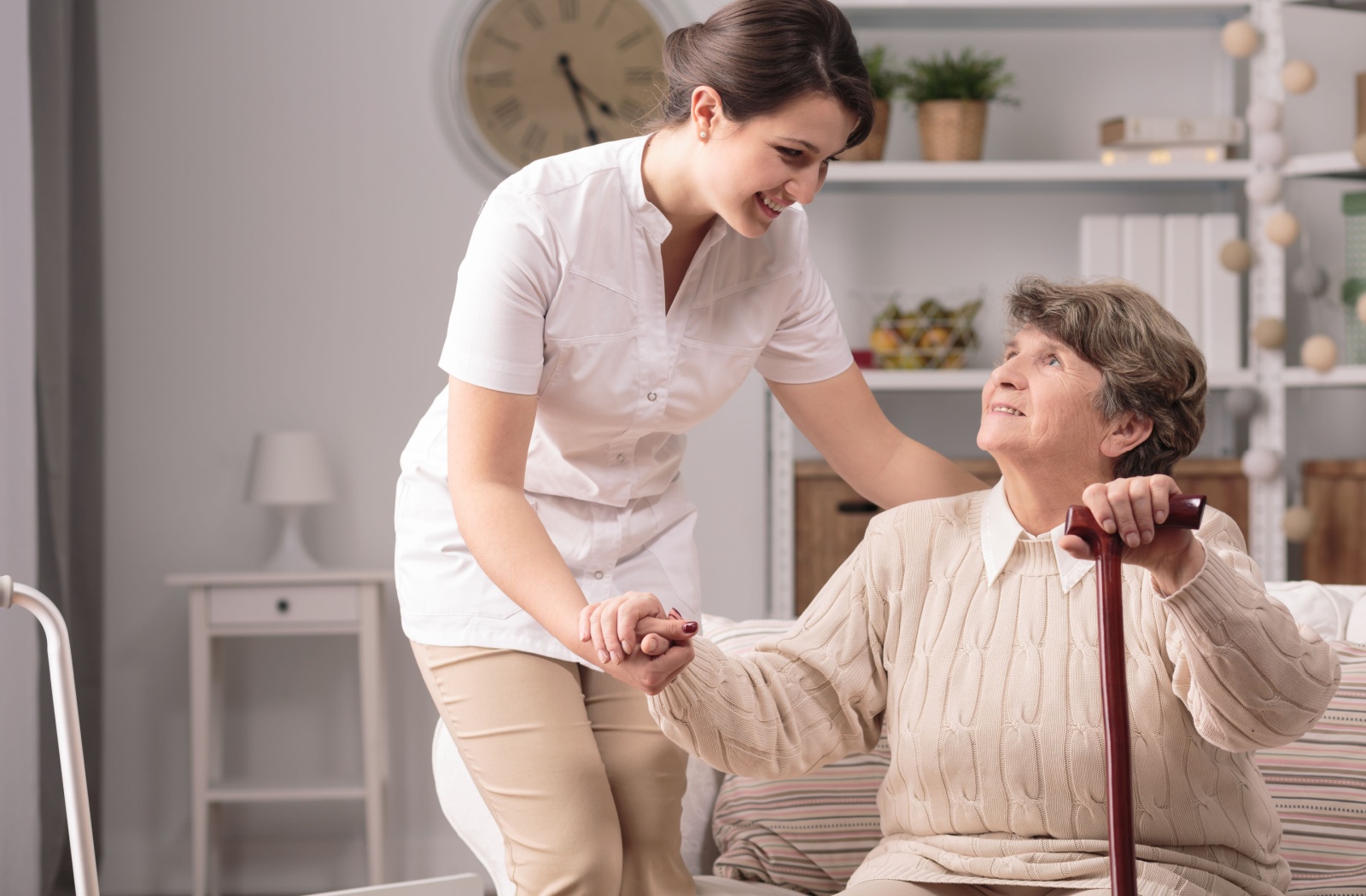 A female caregiver helping a senior woman with a walking stick stand up from the couch while holding her to offer additional support.