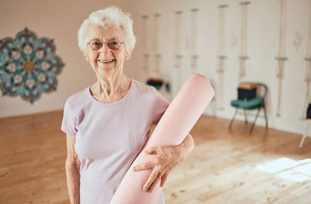 An older adult holding a pink yoga mat smiles at the camera after a balance exercise class in a community space
