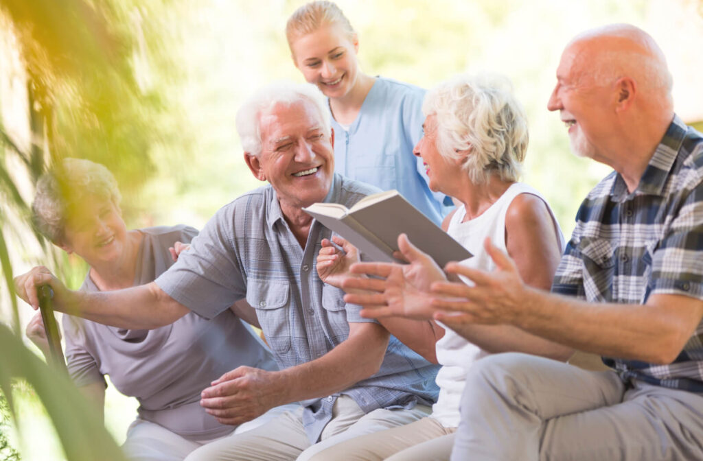 A group of older adults in assisted living laugh while sitting together in an outdoor common area