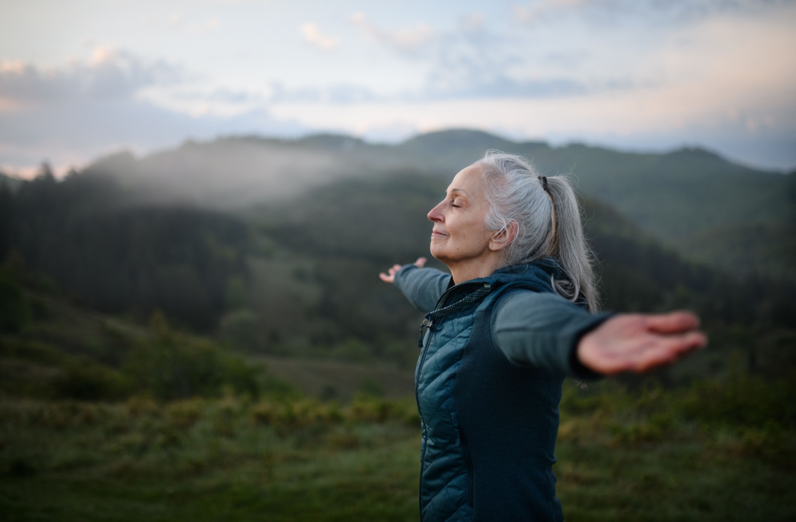 A senior breathes deeply with their arms outstretched while on a morning mountain hike