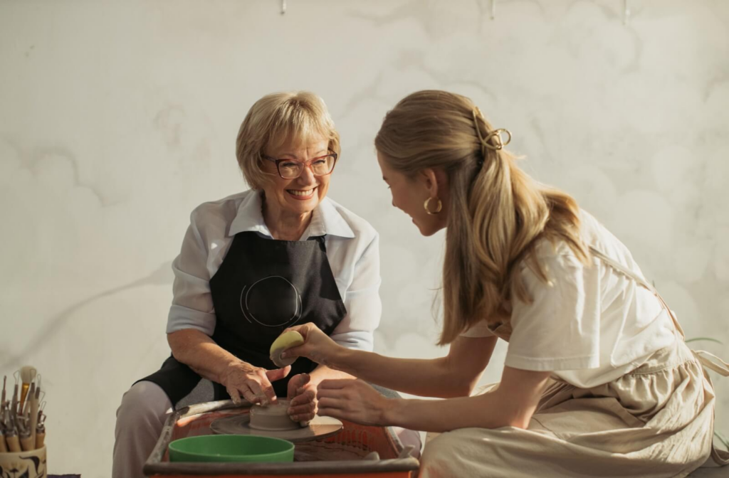 In a bright, clean studio space, a senior smiles at their pottery instructor as they learn how to mold clay to create pottery