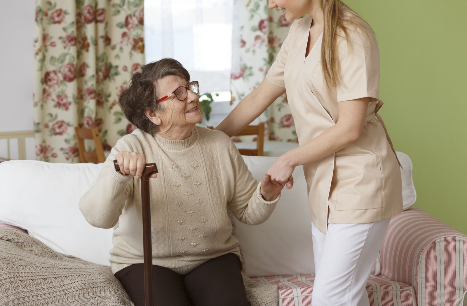 A female caregiver helping an elderly woman stand up from the couch while she uses a walking stick on the other hand.