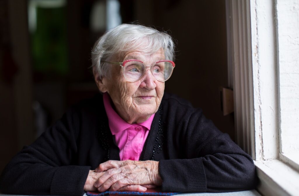 An older, smartly-dressed woman with glasses looks out of a window, pensive and pondering the next stage of life