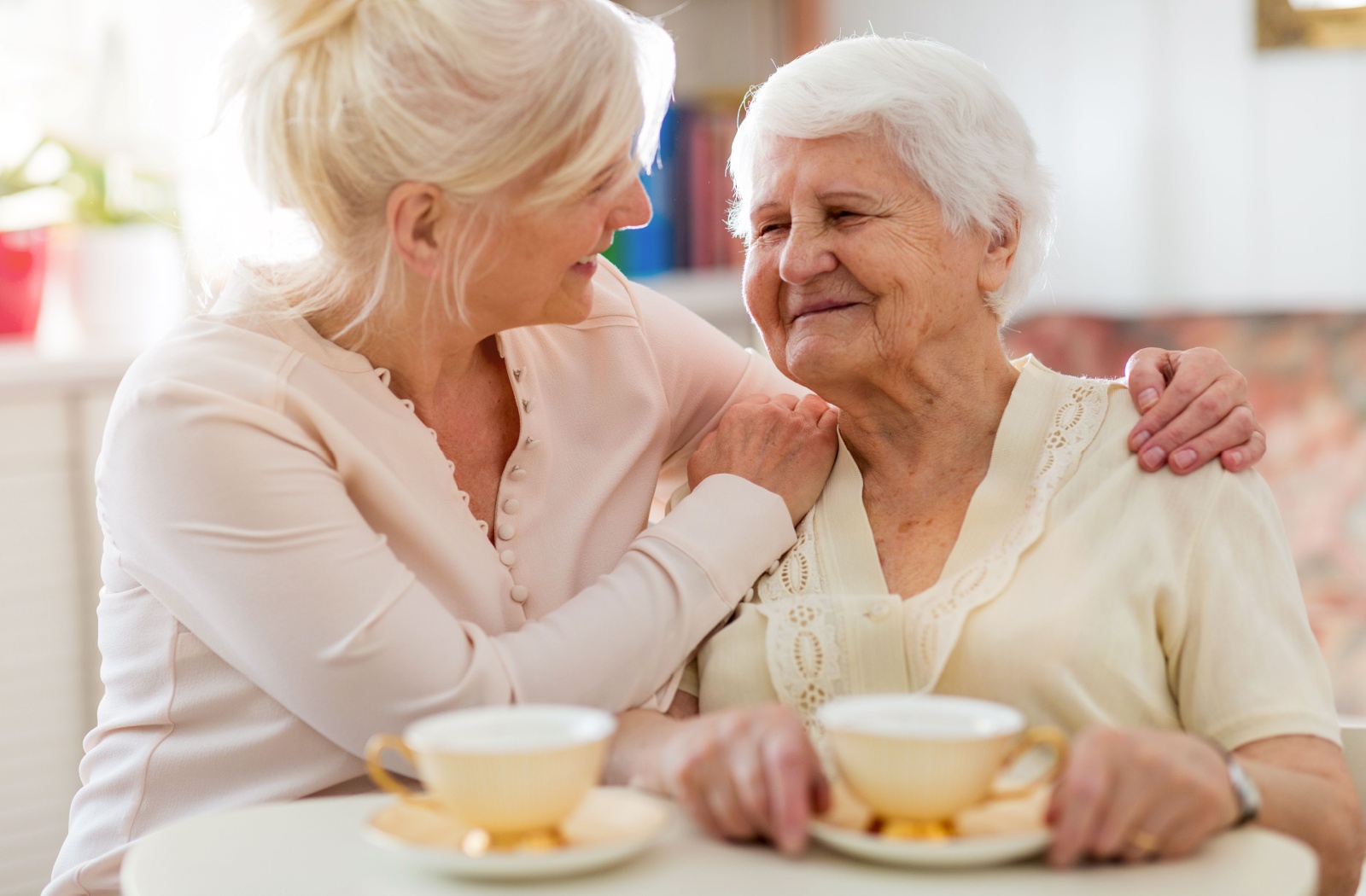 An adult embraces their older parent by the shoulders, smiling at them while the two enjoy cups of tea together
