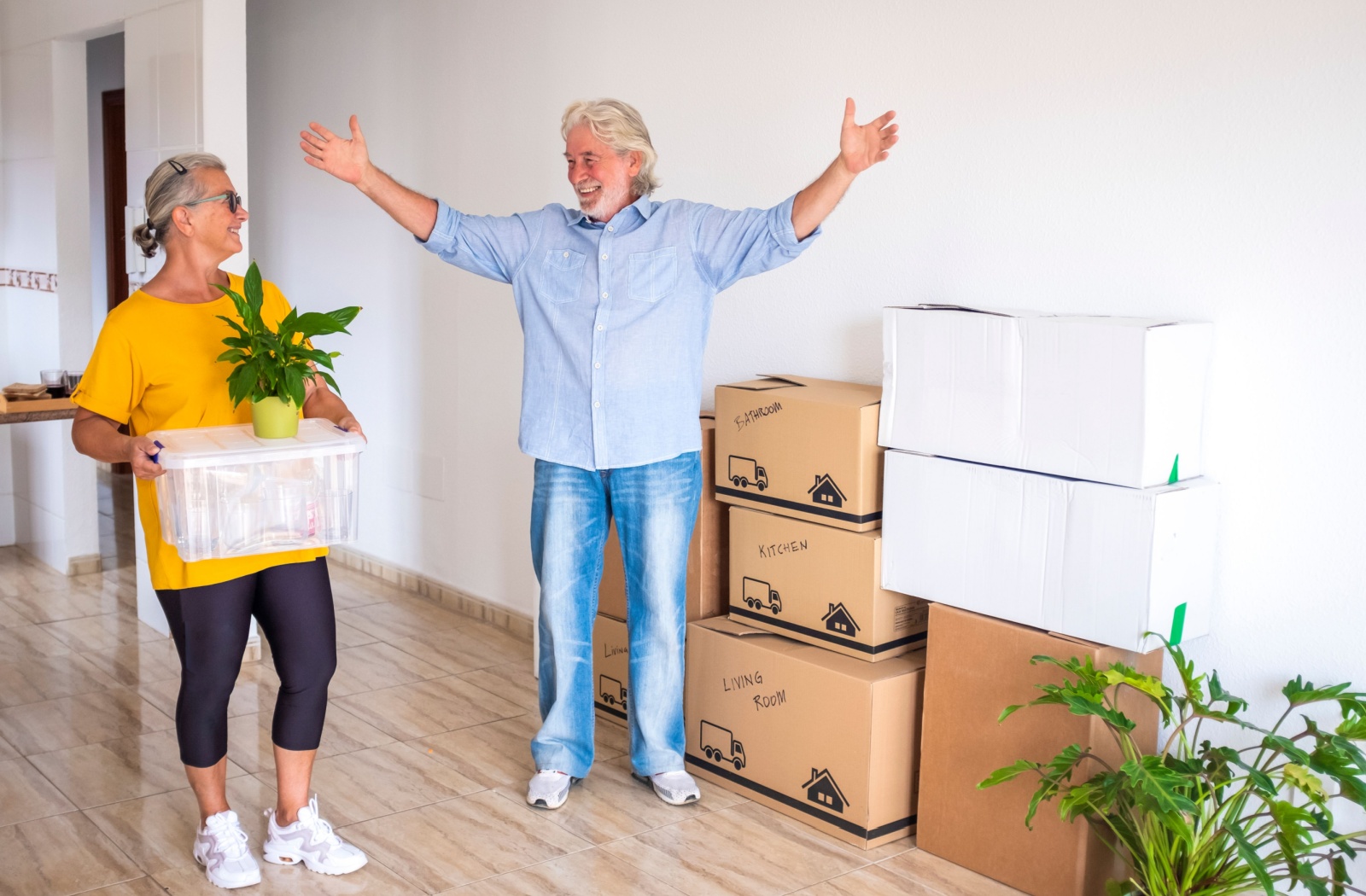 An older adult lifts their arms in celebration in their new home in assisted living, standing in front of a stack of moving boxes beside their laughing spouse